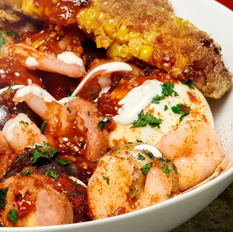 Close-up of shrimp, potatoes, and corn on the cob in a rich red sauce with white drizzle.