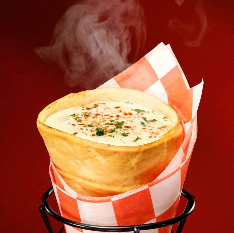 Steaming hot bread cone filled with creamy chowder, garnished with herbs and paprika, in a metal holder.