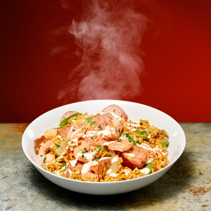 Steaming bowl of jambalaya with shrimp, sausage, rice, and green peppers, drizzled with white sauce.