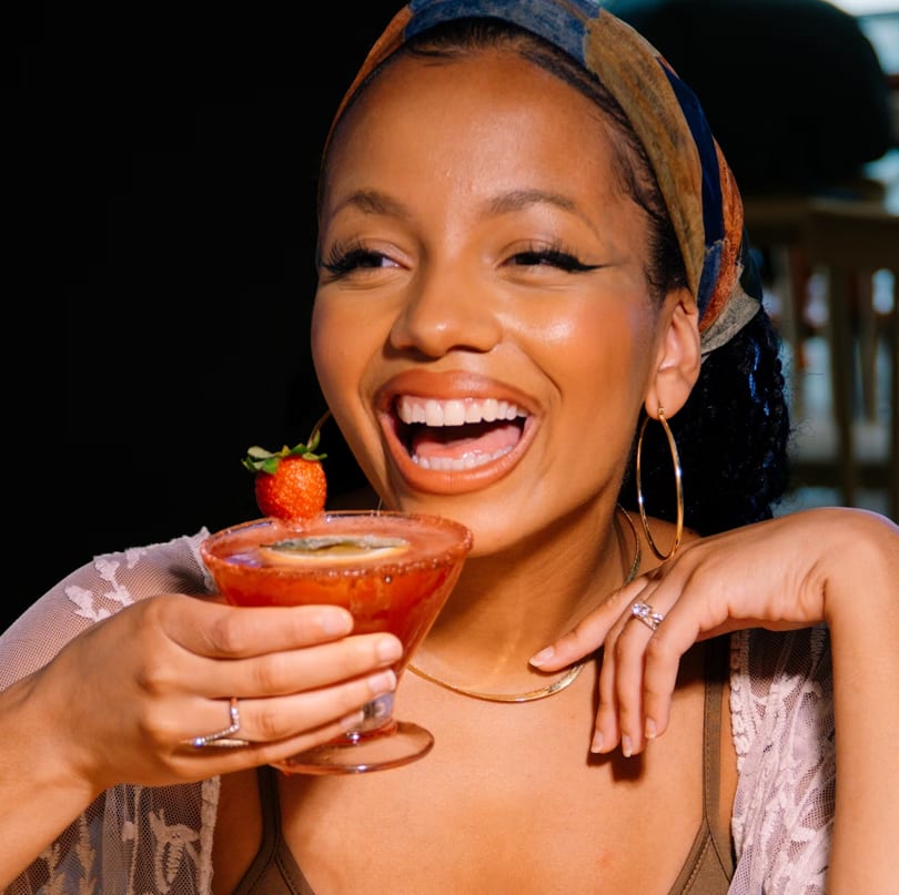 A smiling woman in a headscarf holds a strawberry cocktail, wearing hoop earrings and rings.